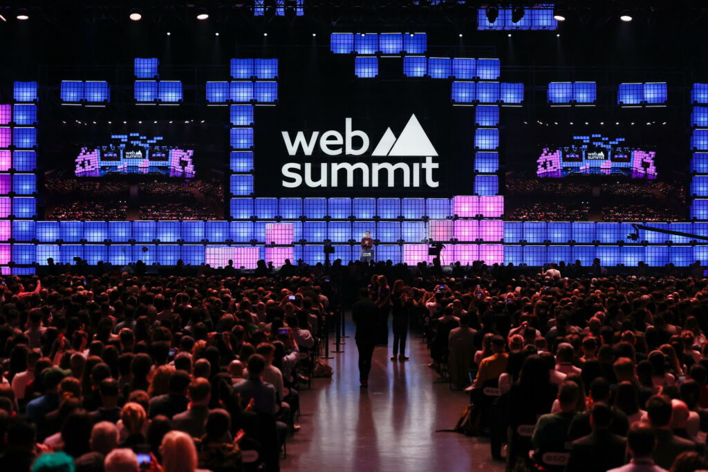 Suuressa hämärässä salissa yleisö istuu paikoillaan ja joku puhuu lavalla. Lavan takana on Web Summit Lissabon -tapahtuman logo.