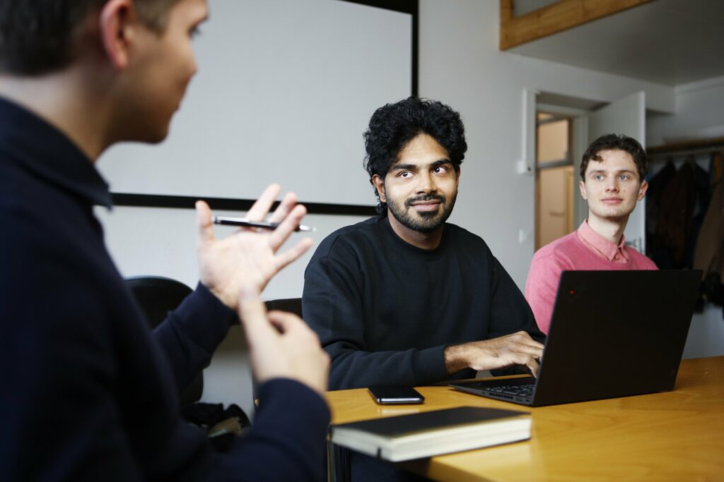 Kolme ihmistä istuu saman pöydän ääressä. Yksi heistä elehtii käsillään, ja toinen katsoo häntä ja näpyttelee kannettavan tietokoneen näppäimistöä.
