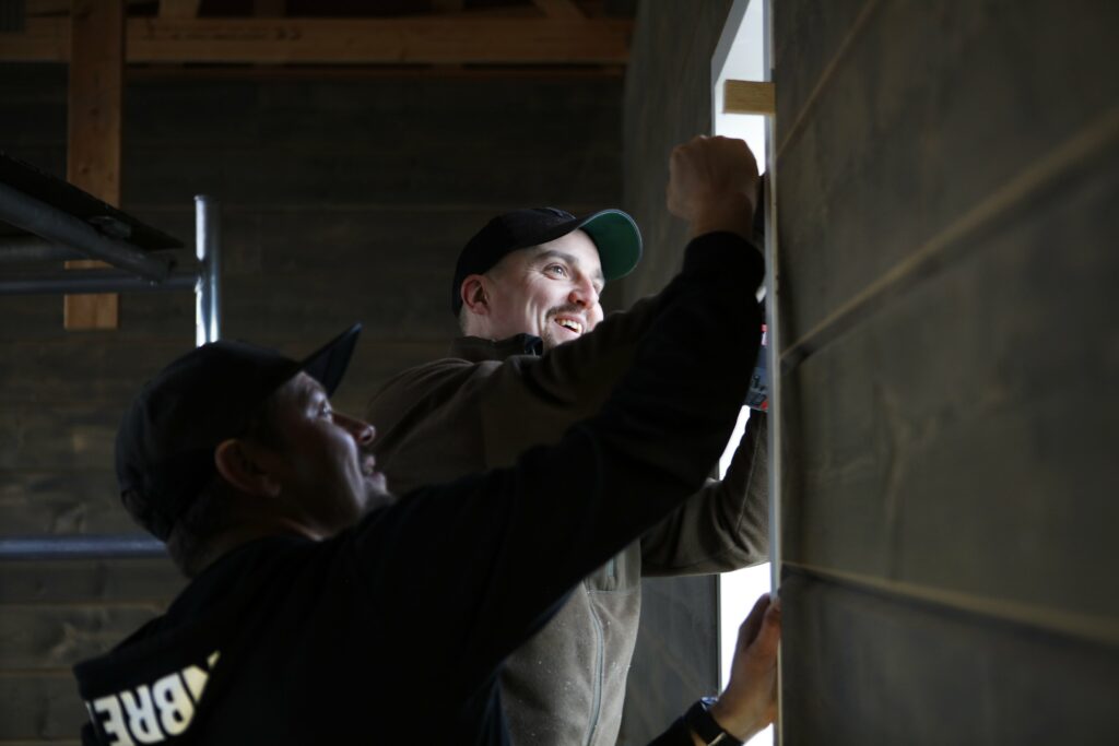 Rakentajat asentavat yhdessä jotain keskeneräisen ikkunan karmiin rakennuksen sisällä.