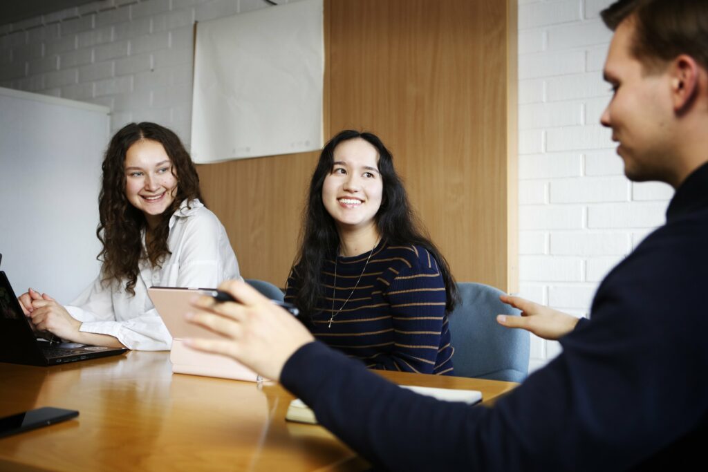 Kaksi hymyilevää naista istuu pöydän ääressä vierekkäin ja katsoo pöydän päässä istuvaa kolmatta henkilöä, joka puhuu ja elehtii käsillään.